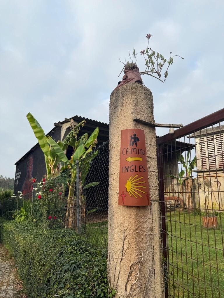 A fencepost showing text "Camino Ingles" is in the foreground, with green foliage and a painted, three-wall structure in the back.