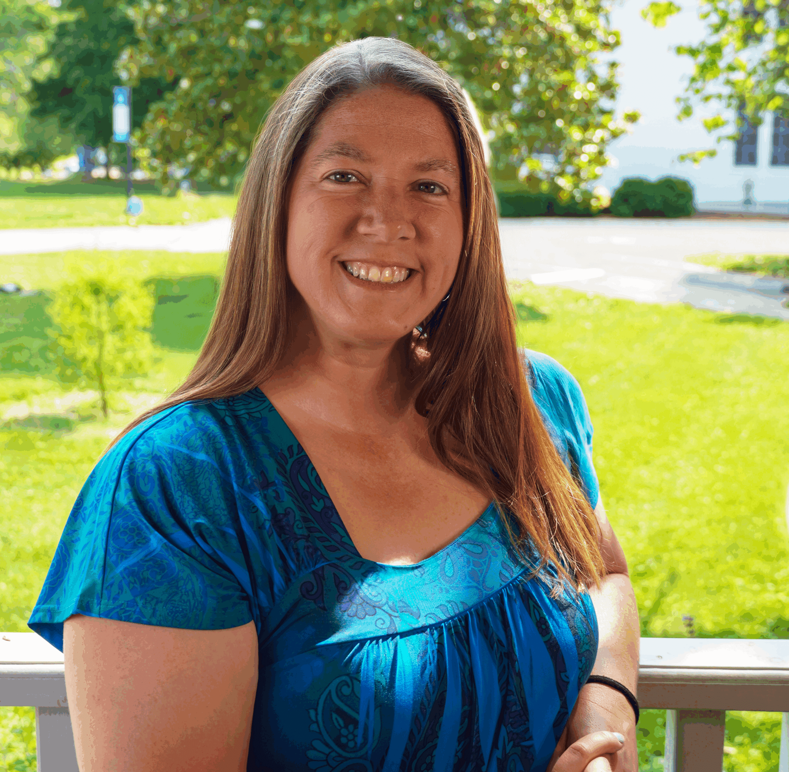 A photo of Regional Admissions Manager for the Southwest Virginia region, Jacinda Pensley, standing outside amongst green grass and trees, leaning against a white railing, wearing a blue short sleeved shirt, smiling.